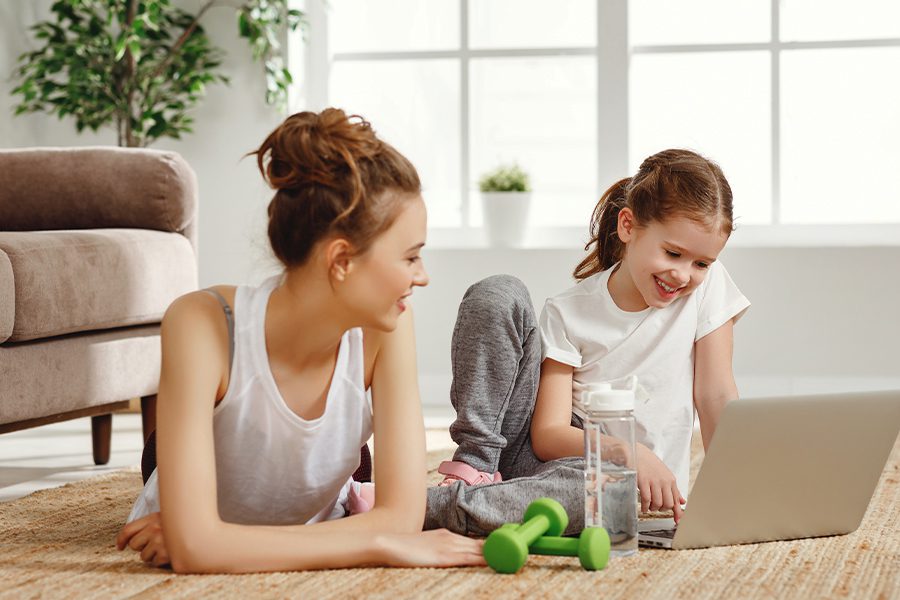 Employee Benefits - Cheerful Mom and Daughter Using Laptop While Exercising at Home